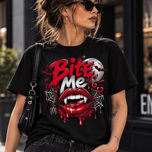 Pode incluir: Uma t-shirt preta de manga curta com uma estampa gráfica de lábios vermelhos com presas de vampiro brancas, líquido vermelho a escorrer e o texto "Bite Me" em vermelho e branco. O design inclui uma lua branca, teias de aranha, pequenos corações vermelhos e morcegos.