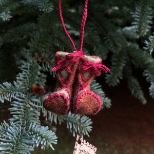 May include: A miniature felt cowboy boot Christmas ornament in shades of brown, green, and red, hanging from a red string on a pine tree branch. The ornament has intricate stitching details and is suspended in front of a blurred green background.