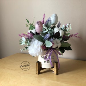 May include: A white ceramic planter with the text "Flowers & Jardin" on the side, filled with a floral arrangement of purple lavender, white cotton, and green eucalyptus. The planter is on a wooden stand and decorated with speckled pastel Easter eggs.