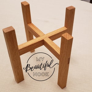 May include: Wooden plant stand with four legs and a cross-shaped design. The stand is made of light brown wood and has a natural finish.
