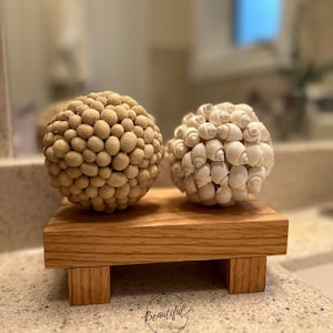 May include: Two decorative spheres made from natural materials. The sphere on the left is made of small, brown, round objects. The sphere on the right is made of white seashells. Both spheres are sitting on a wooden stand with four legs.