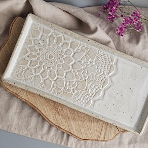 Könnte beinhalten: Ein rechteckiges Keramik-Serviertablett mit einem erhabenen, komplizierten Spitzendesign auf einer Hälfte. Das Tablett ist cremefarben mit gesprenkelten Details und liegt auf einem Holzbrett. Ein Zweig lila Blumen befindet sich im Hintergrund.