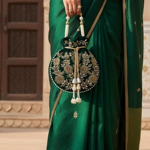 May include: A woman in a dark green sari holds a matching round handbag. The bag is black with gold floral embroidery and white beaded accents. The sari has gold trim and subtle gold embroidery.