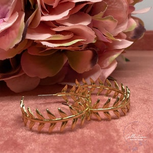 May include: Gold hoop earrings with a leaf design. The earrings are on a pink background.