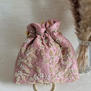 May include: A dusty rose drawstring pouch bag featuring intricate gold thread embroidery and small white pearl bead accents. The bag has a gathered top closure with gold-toned hardware and a circular gold metal handle at the base.