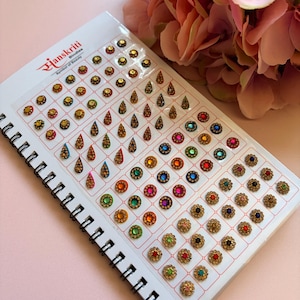 May include: A spiral-bound display card labelled Hanskriti Bindies featuring an assortment of adhesive face stickers in various shapes, including small circles, teardrops, and floral designs, accented with colourful rhinestones and gold-tone metal.