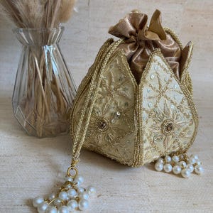 May include: A gold-coloured, beaded pouch with a drawstring closure. The pouch features detailed embroidery, sequins, and a beaded tassel. The bag is approximately 15 cm high and 13 cm wide, with a pearl-like tassel.