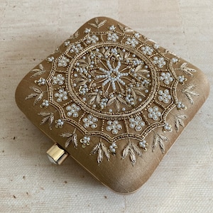 May include: A square-shaped clutch bag with a gold fabric exterior. The clutch is embellished with a circular design of gold and white beads and sequins.