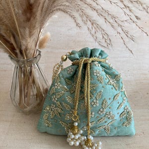 May include: A teal drawstring pouch with gold floral embroidery and white pearl detailing. The pouch features a gold drawstring and a tassel of white pearls. A vase of dried plants is in the background.