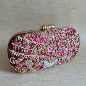 May include: A pink, rectangular clutch bag with detailed floral embroidery. It has gold-coloured accents, white beads, and a gold clasp. The design incorporates floral and leaf patterns.