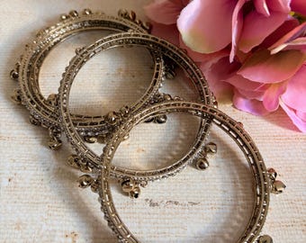 Może przedstawiać: Trzy złote metalowe bransoletki typu bangle o okrągłym kształcie, ozdobione osadzonymi przezroczystymi kryształkami oraz małymi dzwoneczkami przymocowanymi w regularnych odstępach wzdłuż zewnętrznej krawędzi każdej z bransoletek.