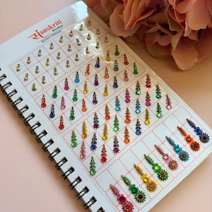 May include: A spiral-bound display card labelled Hanskriti Symbol of Beauty featuring an assortment of adhesive bindis in various shapes, sizes, and colours including red, blue, green, yellow, pink, and orange, arranged in rows and columns.