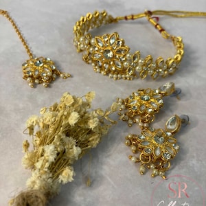 May include: A gold-tone jewellery set with white stones and pearls. The set includes a choker necklace, a maang tikka, and a pair of earrings. The jewellery is arranged on a white background with dried flowers.