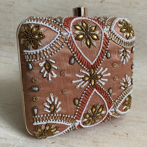 Könnte beinhalten: Eine kleine, quadratische Clutch mit einem goldenen Verschluss. Die Clutch ist aus einem pfirsichfarbenen Stoff gefertigt und mit aufwendigen Stickereien in Gold und Weiß verziert. Die Stickereien zeigen florale und geometrische Muster.