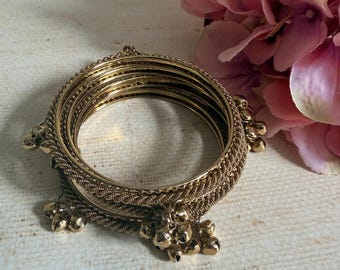 Może przedstawiać: Zestaw złotych metalowych bransoletek typu bangle o teksturowanej, plecionej powierzchni. Bransoletki są ozdobione małymi, zwisającymi złotymi dzwoneczkami przymocowanymi w odstępach wzdłuż zewnętrznych krawędzi okrągłych metalowych obręczy.