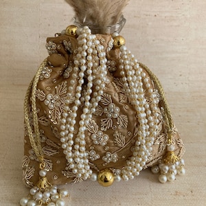 May include: A gold embroidered drawstring bag with pearl embellishments and gold tassels. The bag is decorated with floral patterns and has a beaded drawstring closure.