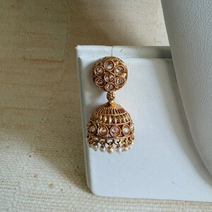 May include: A gold-toned jhumka earring featuring a circular stud top with embedded clear stones. The bell-shaped drop is decorated with intricate metal leaf patterns, additional clear stones, and a row of small white beads hanging from the bottom edge.