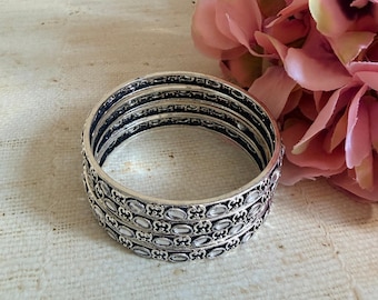 Może przedstawiać: Zestaw trzech srebrzystych metalowych bransoletek typu bangle z powtarzającym się owalnym wzorem i ciemnymi wgłębieniami. Bransoletki są ułożone jedna na drugiej, prezentując metaliczne wykończenie ze skomplikowanymi tłoczonymi teksturami.