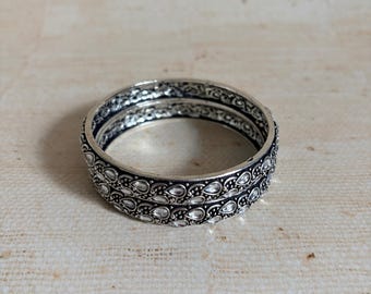 Może przedstawiać: Para srebrzystych bransoletek typu bangle z misternym tłoczonym wzorem z powtarzającymi się elementami w kształcie łezek. Powierzchnia posiada ciemne, oksydowane wykończenie we wgłębieniach, które podkreśla szczegółowy wzór metaloplastyki.