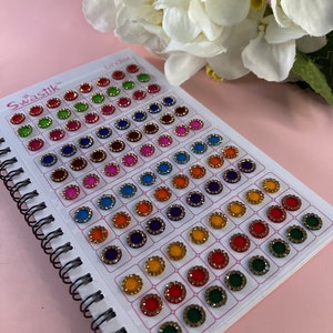 May include: A sheet of colourful round stickers with gold trim. The stickers are arranged in rows and columns on a white sheet with the word "Swastik" printed in the top left corner. The sheet is bound with a black spiral binding.