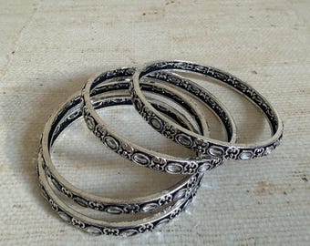 Może przedstawiać: Zestaw trzech srebrzystych metalowych bransoletek typu bangle z powtarzającym się tłoczonym wzorem owalnych kształtów i ozdobnych zawijasów. Bransoletki mają ciemne, oksydowane wykończenie w zagłębieniach, które podkreśla wypukłe detale.