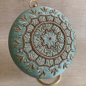 May include: A round, light blue clutch bag with a gold metal clasp and intricate gold embroidery featuring a circular design with white beads.