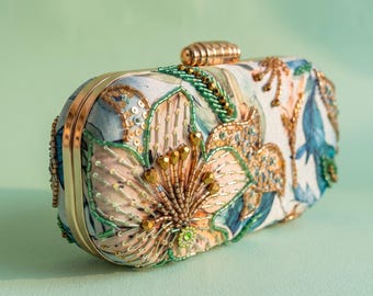 Może przedstawiać: Prostokątna kopertówka z kwiatowym wzorem w odcieniach niebieskiego, brzoskwiniowego i białego. Powierzchnia ozdobiona jest złotymi, zielonymi i przezroczystymi koralikami oraz cekinami. Posiada złotą metalową ramkę i żebrowane zapięcie w kolorze złota.