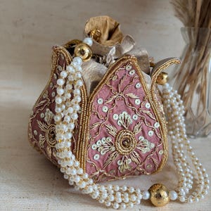 May include: A dusty rose drawstring pouch bag featuring intricate gold embroidery, metallic sequins, and beadwork. The bag has a structured petal-like shape and is adorned with long, multi-strand white pearl bead handles with gold-toned accents.
