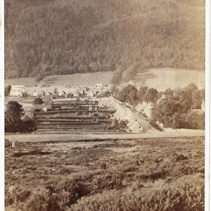 Op de afbeelding: Een zwart-witfoto van een klein stadje dat in een vallei ligt, met een berg op de achtergrond. Het stadje is omgeven door bomen en velden.