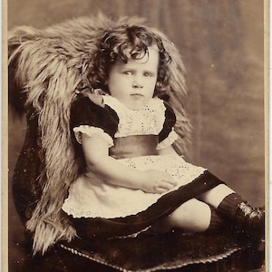 May include: A young girl with curly hair sits on a chair with a fur throw. She is wearing a white dress with lace trim and a dark sash.