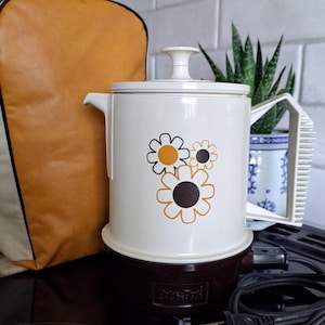 May include: An off-white electric percolator with a brown base and a black power cord. The body features a graphic print of three stylized flowers in shades of orange, brown, and white. It has a matching off-white lid with a knob and a handle.
