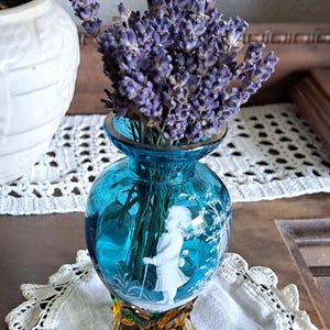 May include: A small, blue glass vase with a white etched design of a girl in a garden. The vase has three amber-colored legs and is filled with dried lavender.