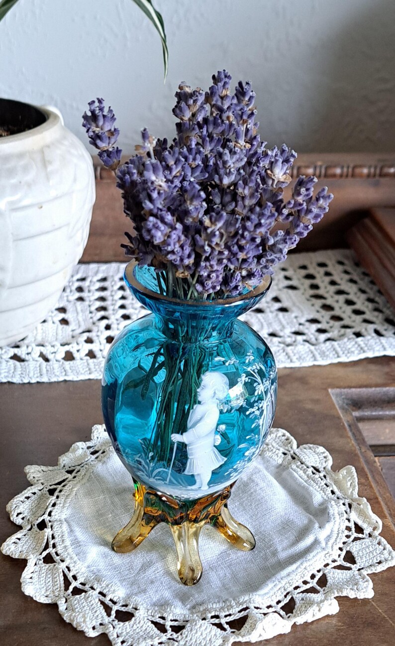 May include: A small, blue glass vase with a white etched design of a girl in a garden. The vase has three amber-colored legs and is filled with dried lavender.