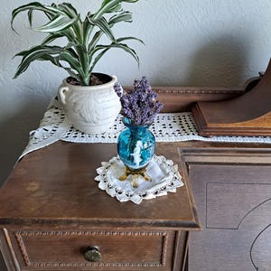 May include: A teal blue glass vase with a white silhouette of a woman and a bouquet of lavender flowers. The vase has a gold base and sits on a white crocheted doily.