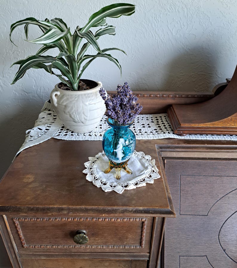 May include: A teal blue glass vase with a white silhouette of a woman and a bouquet of lavender flowers. The vase has a gold base and sits on a white crocheted doily.