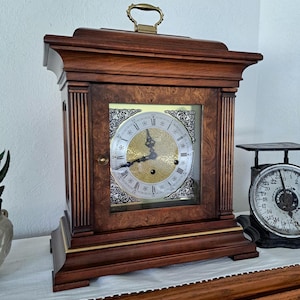 May include: A wooden mantel clock with a dark brown finish and a burl wood front panel. The clock features a gold-toned face with black Roman numerals, ornate silver-colored corner accents, and a brass handle on the top. The clock has a square glass door.