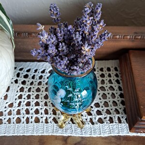 May include: A bouquet of dried lavender flowers in a small, blue glass vase with a gold rim and three gold legs. The vase has a white floral design.