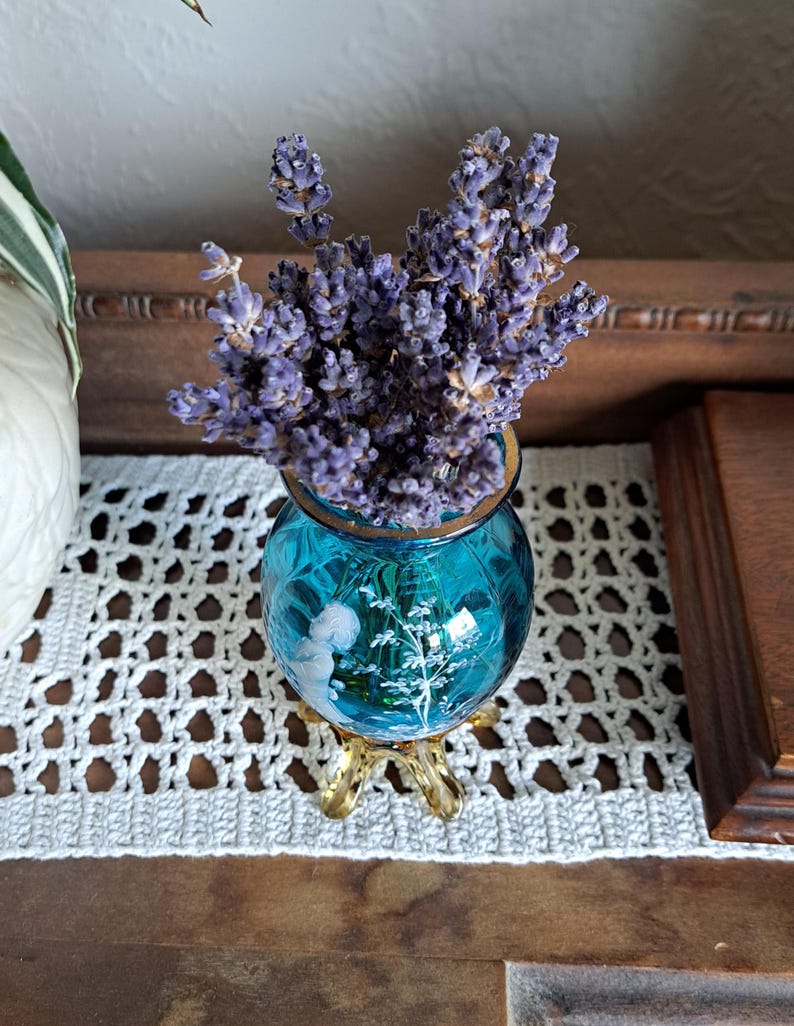 May include: A bouquet of dried lavender flowers in a small, blue glass vase with a gold rim and three gold legs. The vase has a white floral design.