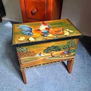 May include: A rectangular wooden stool featuring a hand-painted top surface with a rooster, hen, and chicks. The front panel displays a painted rural landscape with trees, a white fence, and farm buildings, all set against a warm, golden-toned background.