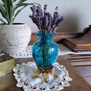 May include: A small, blue glass vase with a bulbous body and three amber-colored legs. The vase is filled with a bouquet of dried lavender.