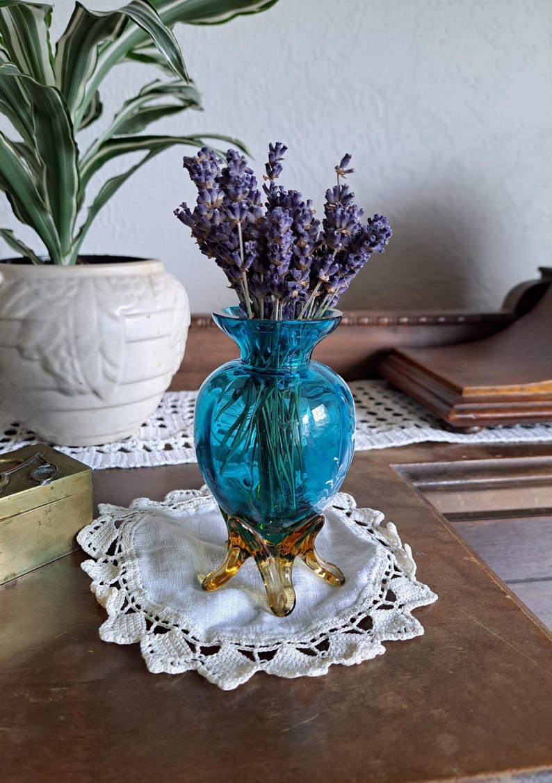 May include: A small, blue glass vase with a bulbous body and three amber-colored legs. The vase is filled with a bouquet of dried lavender.