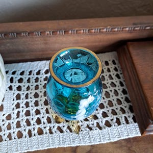May include: A small, blue glass vase with a gold rim and three gold feet. The vase has a white floral design.