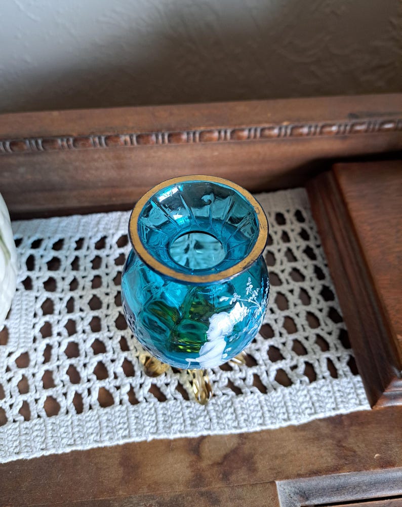 May include: A small, blue glass vase with a gold rim and three gold feet. The vase has a white floral design.