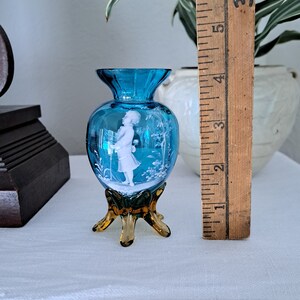 May include: A small, blue glass vase with a white cameo of a girl holding a flower. The vase has a brown, three-legged base.
