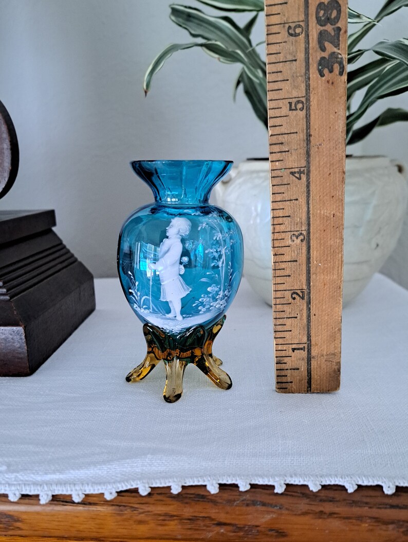 May include: A small, blue glass vase with a white cameo of a girl holding a flower. The vase has a brown, three-legged base.