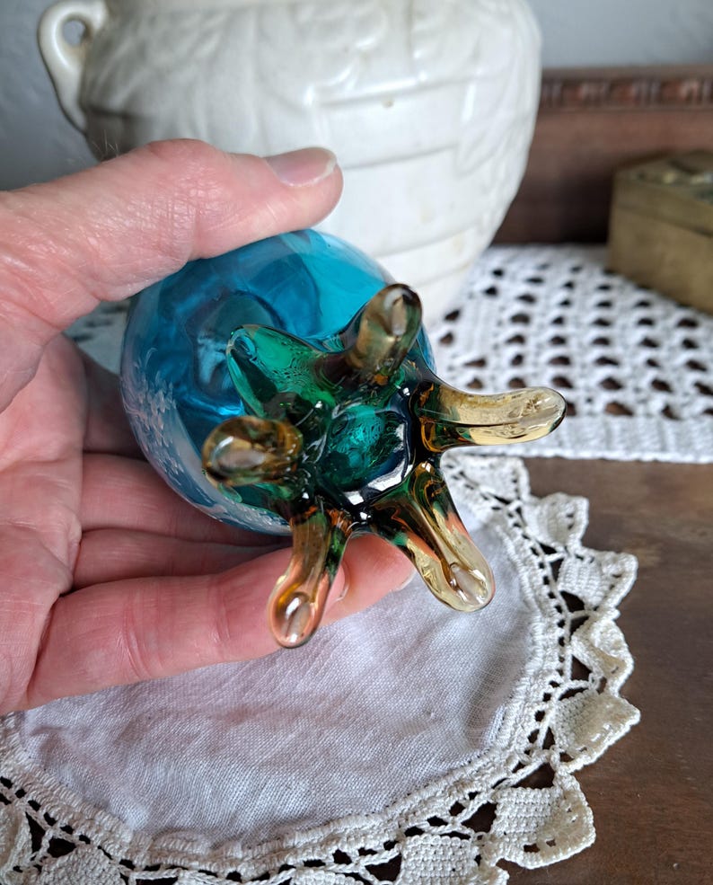 May include: A blue glass vase with a green and brown swirl design. The vase has a unique, spidery base with six legs.