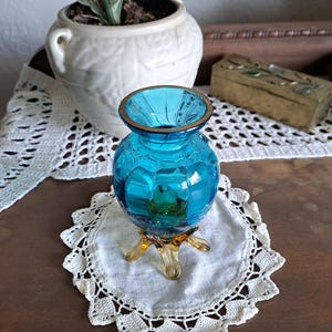 May include: A small, turquoise blue glass vase with a gold rim and three amber-colored feet. The vase is sitting on a white crocheted doily.