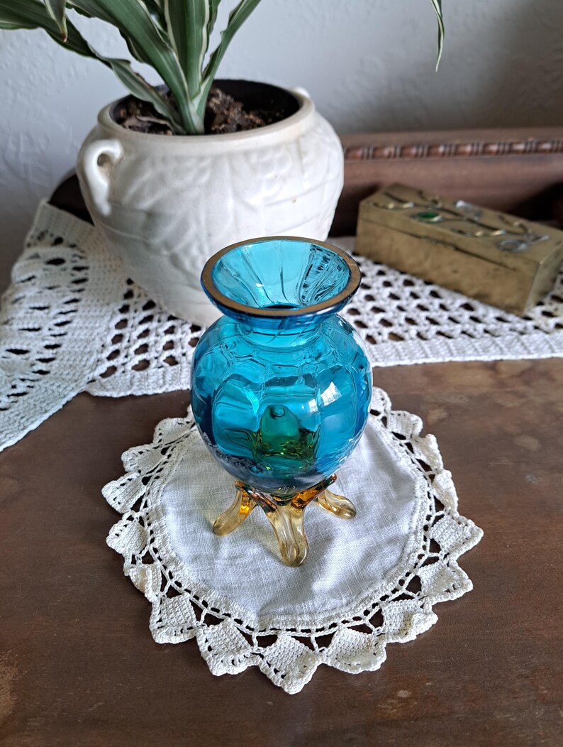 May include: A small, turquoise blue glass vase with a gold rim and three amber-colored feet. The vase is sitting on a white crocheted doily.