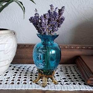 May include: A small, teal blue glass vase with a gold base and a floral design. The vase is filled with dried lavender flowers.