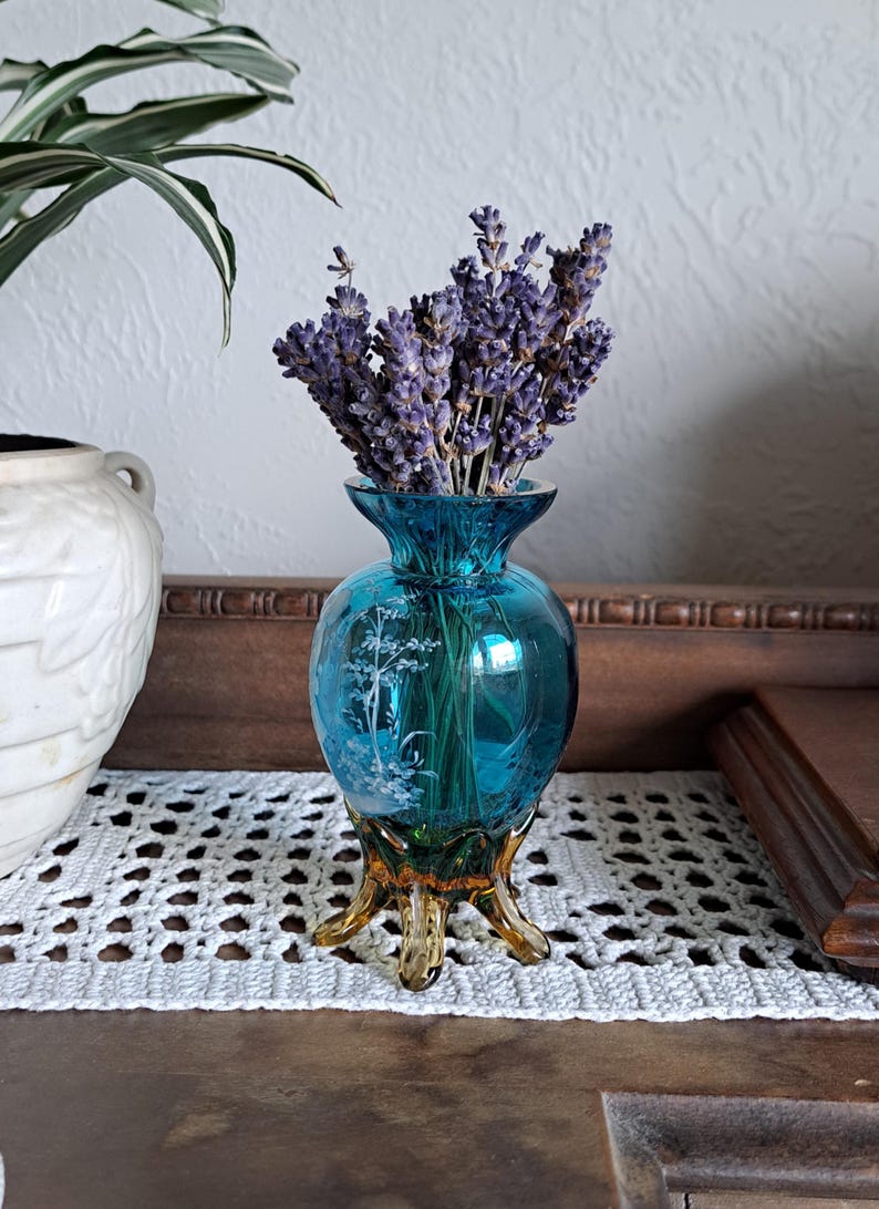 May include: A small, teal blue glass vase with a gold base and a floral design. The vase is filled with dried lavender flowers.